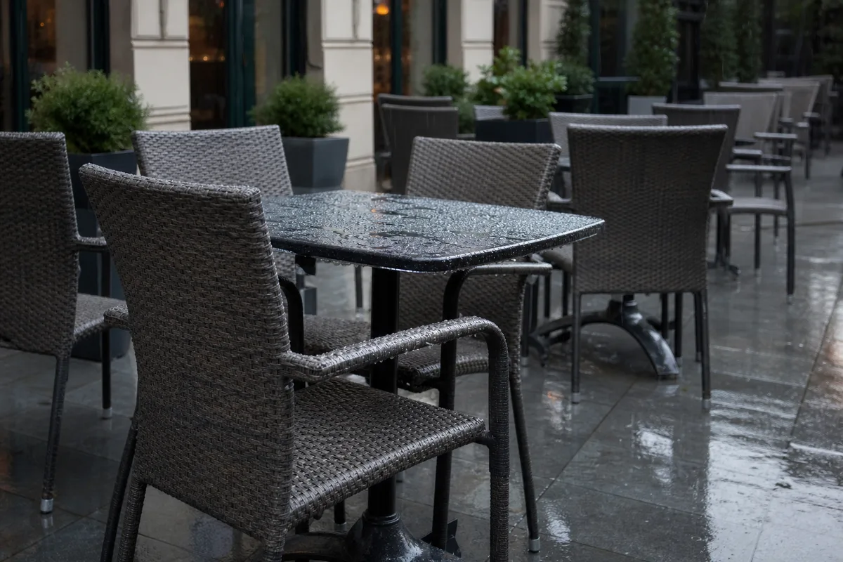 Terrasse de restaurant sous la pluie fine, gouttes séchant sur des chaises en résine tressée